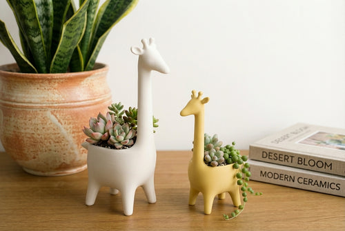 Two giraffe-shaped planters with succulents on a wooden surface next to a potted plant and books.